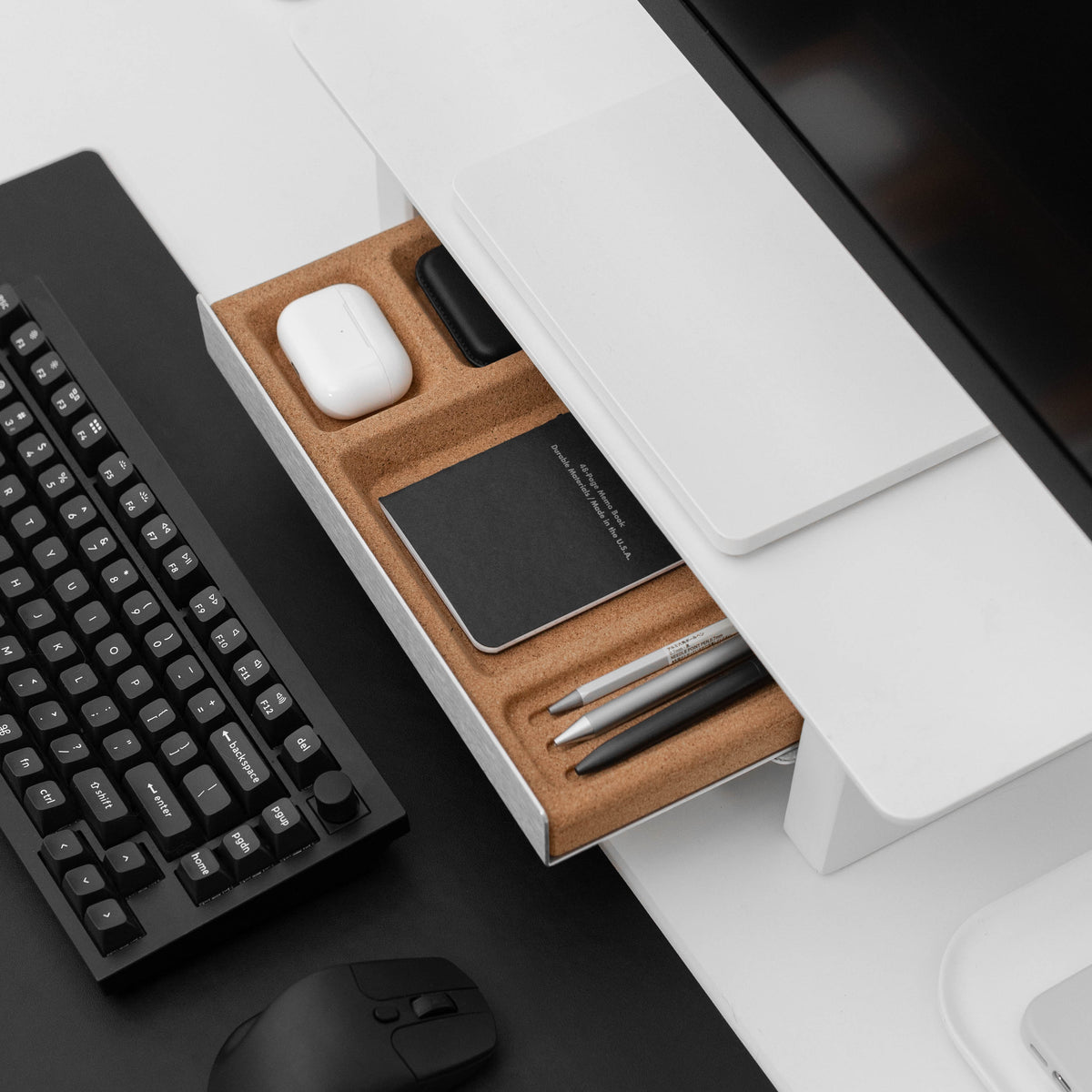 A modern desk organizer drawer with a cork interior, shown open on a white desk next to a black keyboard, mouse, and computer monitor.